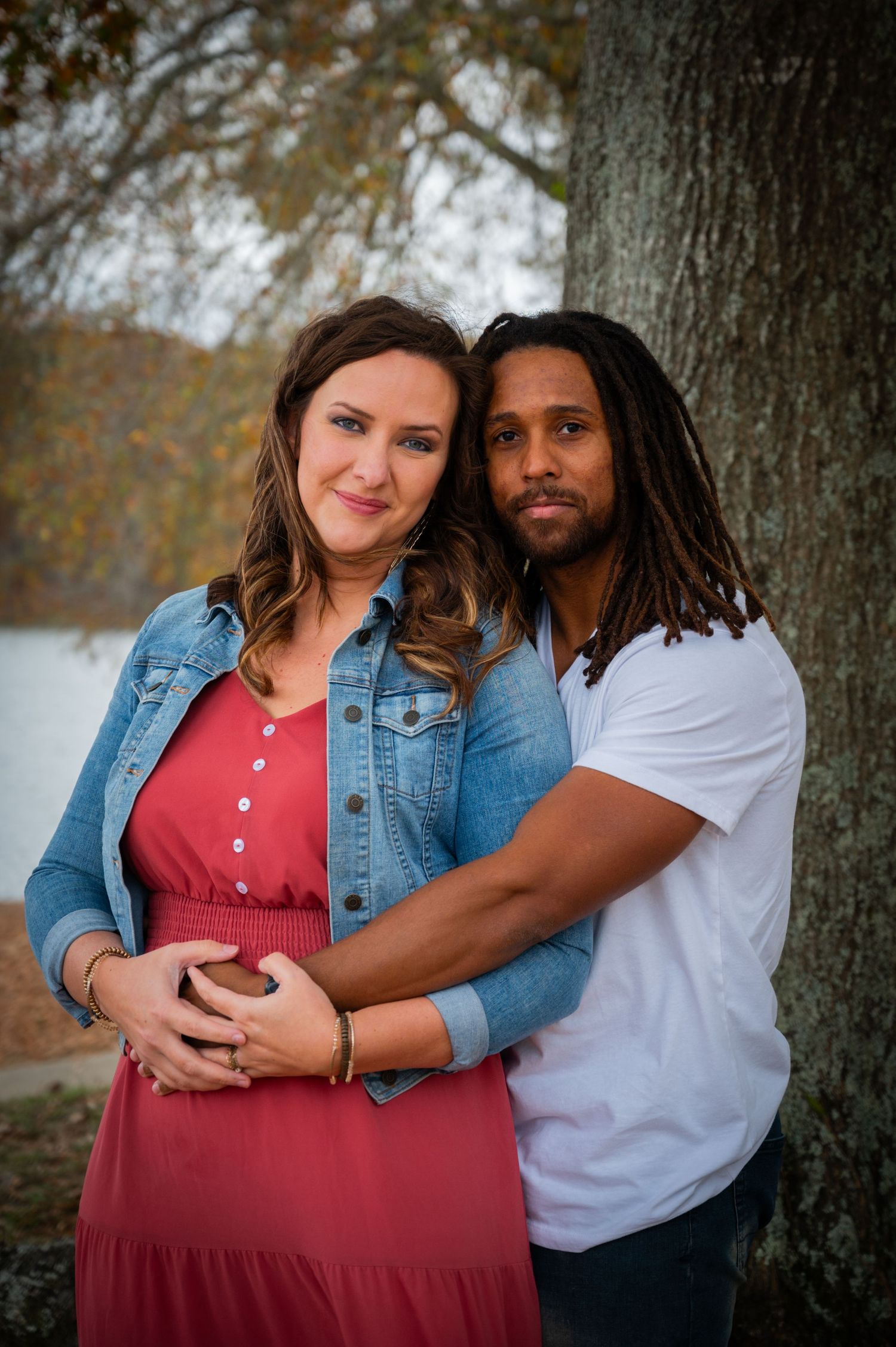 Romantic couple poses for engagement photos in casual attire against natural woodland setting.