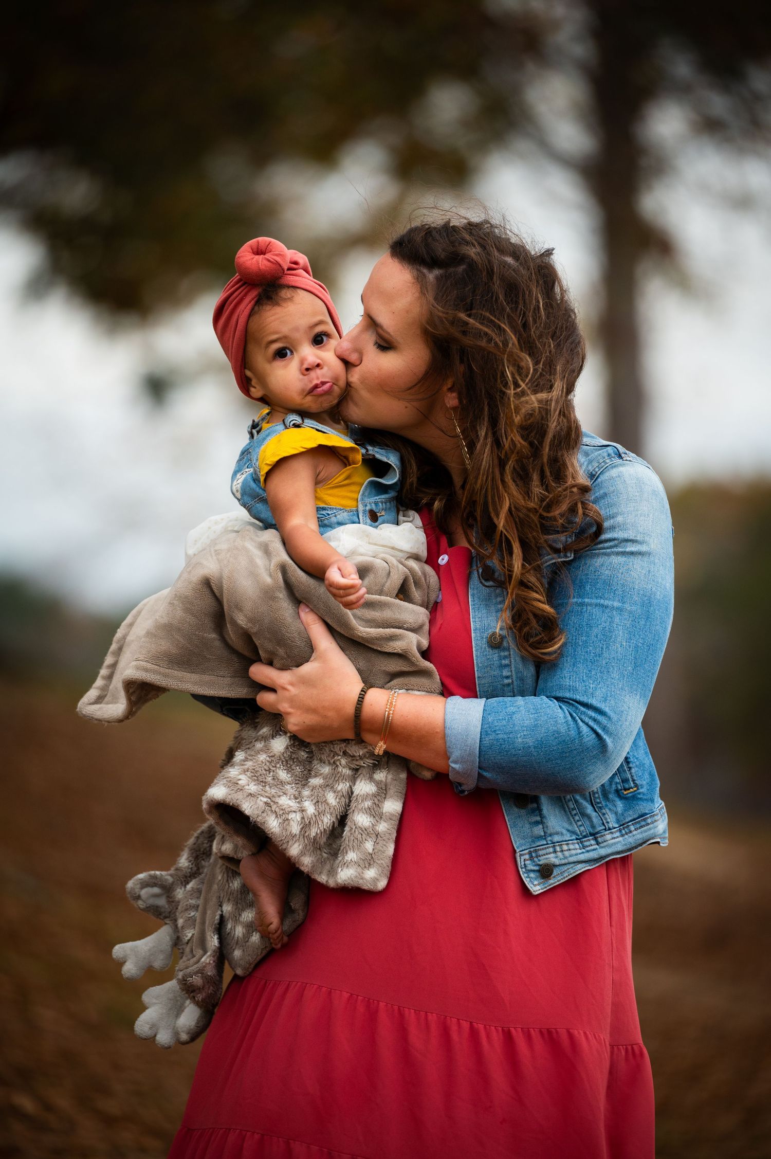 Mother in denim jacket and red dress shares tender moment with baby during outdoor photoshoot.