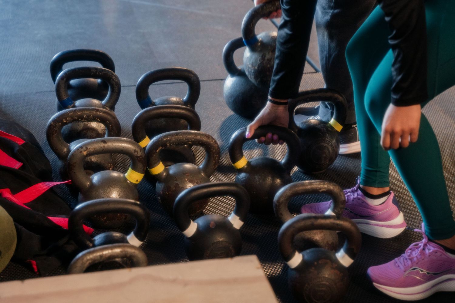 Collection of kettlebells lined up on the floor of a gym with someone's feet visible in frame.