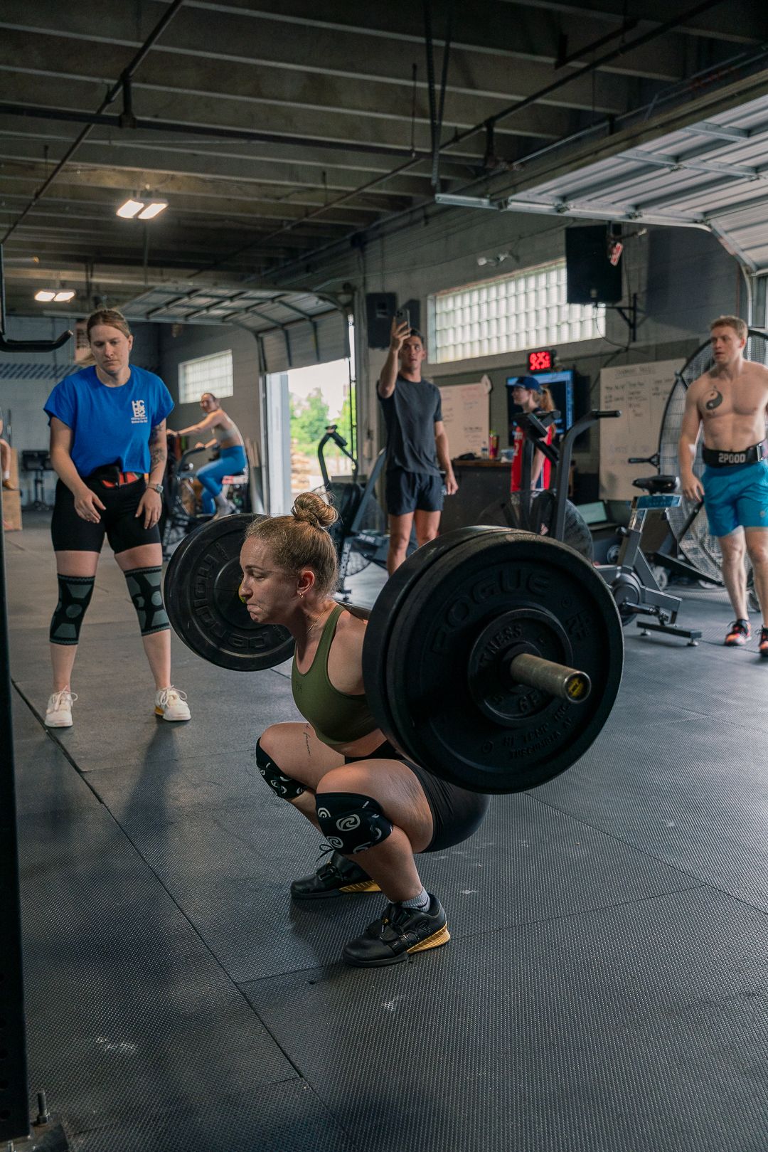 Athletes training at a CrossFit gym with one person performing a heavy barbell squat while others watch and prepare.