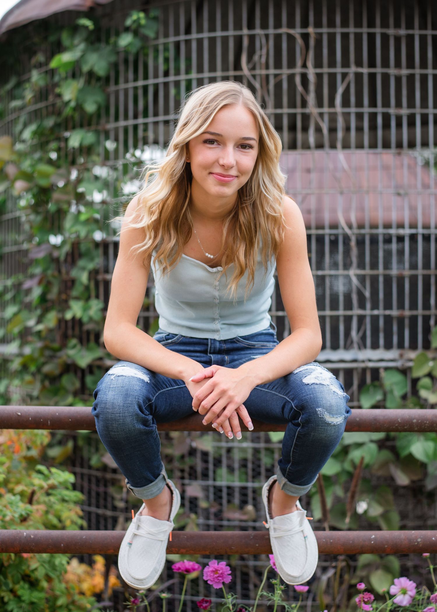 A person in ripped jeans and white sneakers sits casually on a metal railing near flowers and vines.