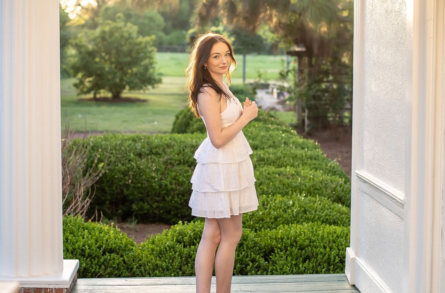 Young woman in a white tiered sundress poses outdoors in a garden setting with architectural columns.
