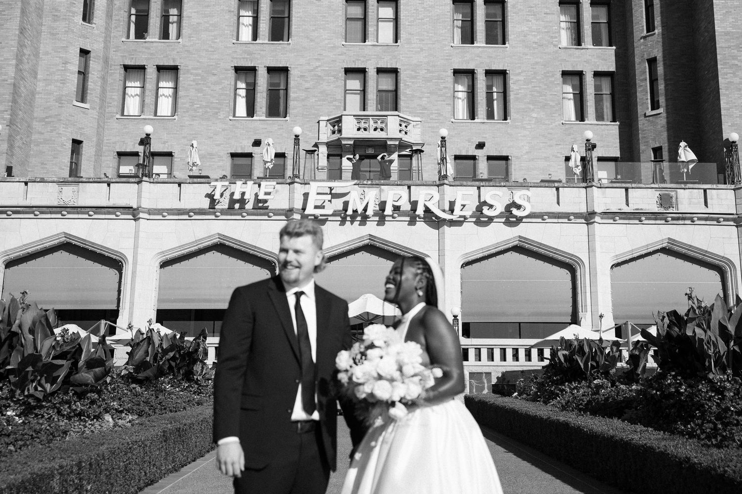Bride holding bouquet gazes at groom surrounded by blurred moving wedding guests in black and white.