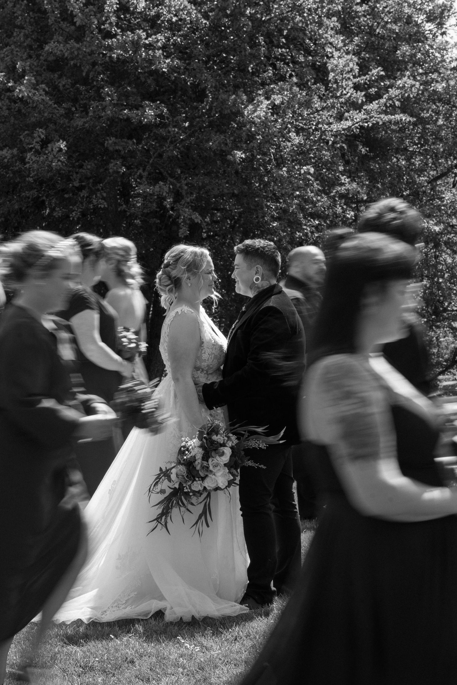 Bride holding bouquet gazes at groom surrounded by blurred moving wedding guests in black and white.