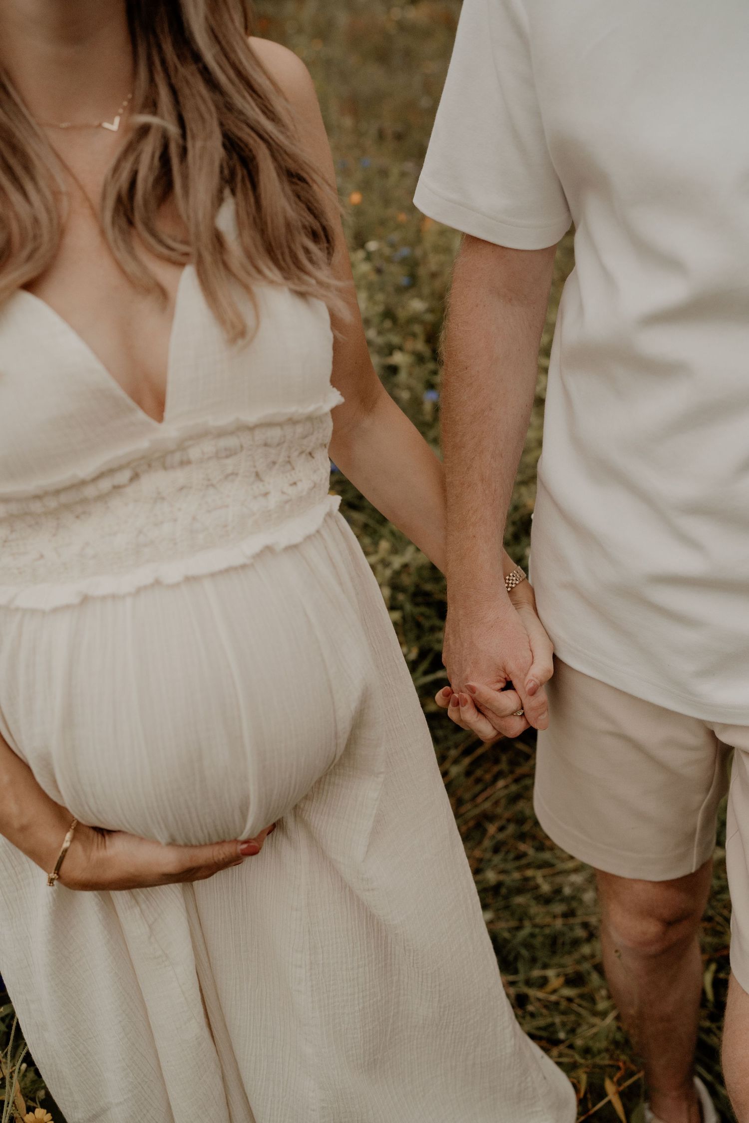 Een stel in het wit houdt hand in hand met een vrouw die haar zwangere buik in een zwierige zwangerschapsjurk vasthoudt, buiten.