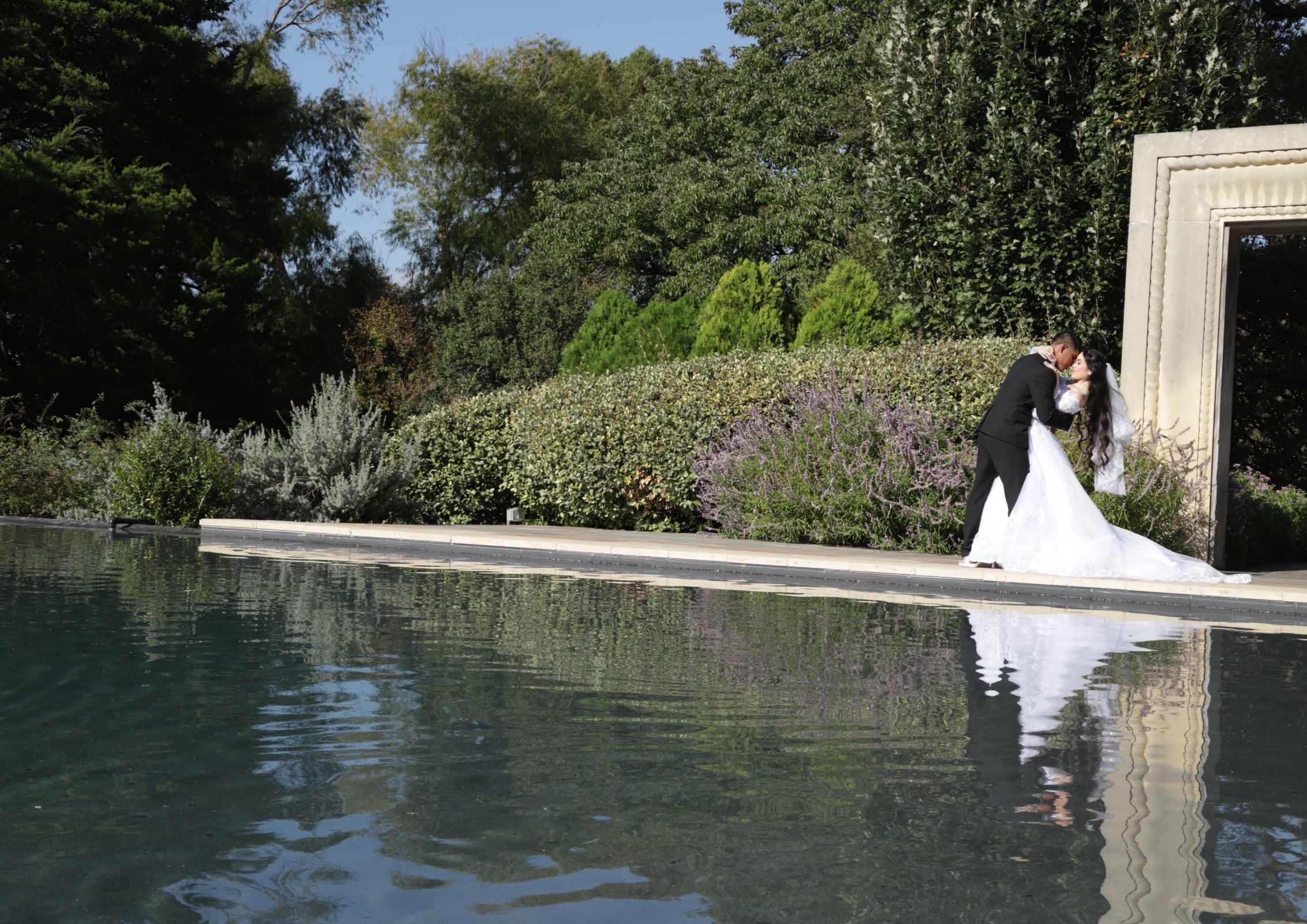 Romantic wedding dress and veil reflection captured in a serene pond with garden backdrop.