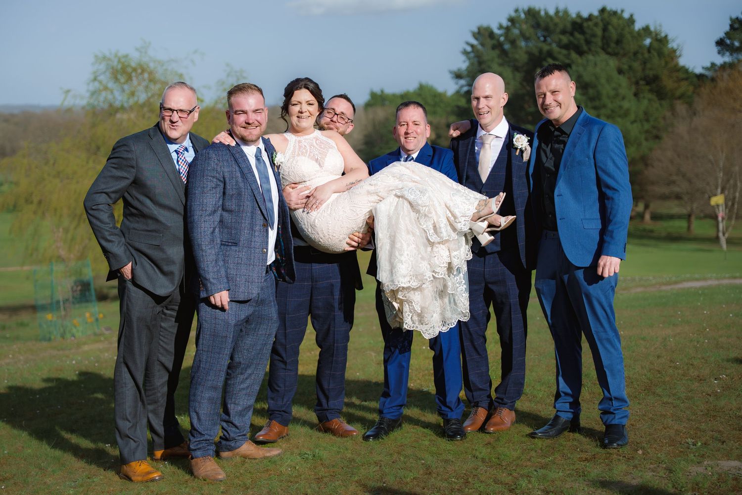 A group of seven people in formal attire stand together outdoors on a grassy hill during a celebration event.