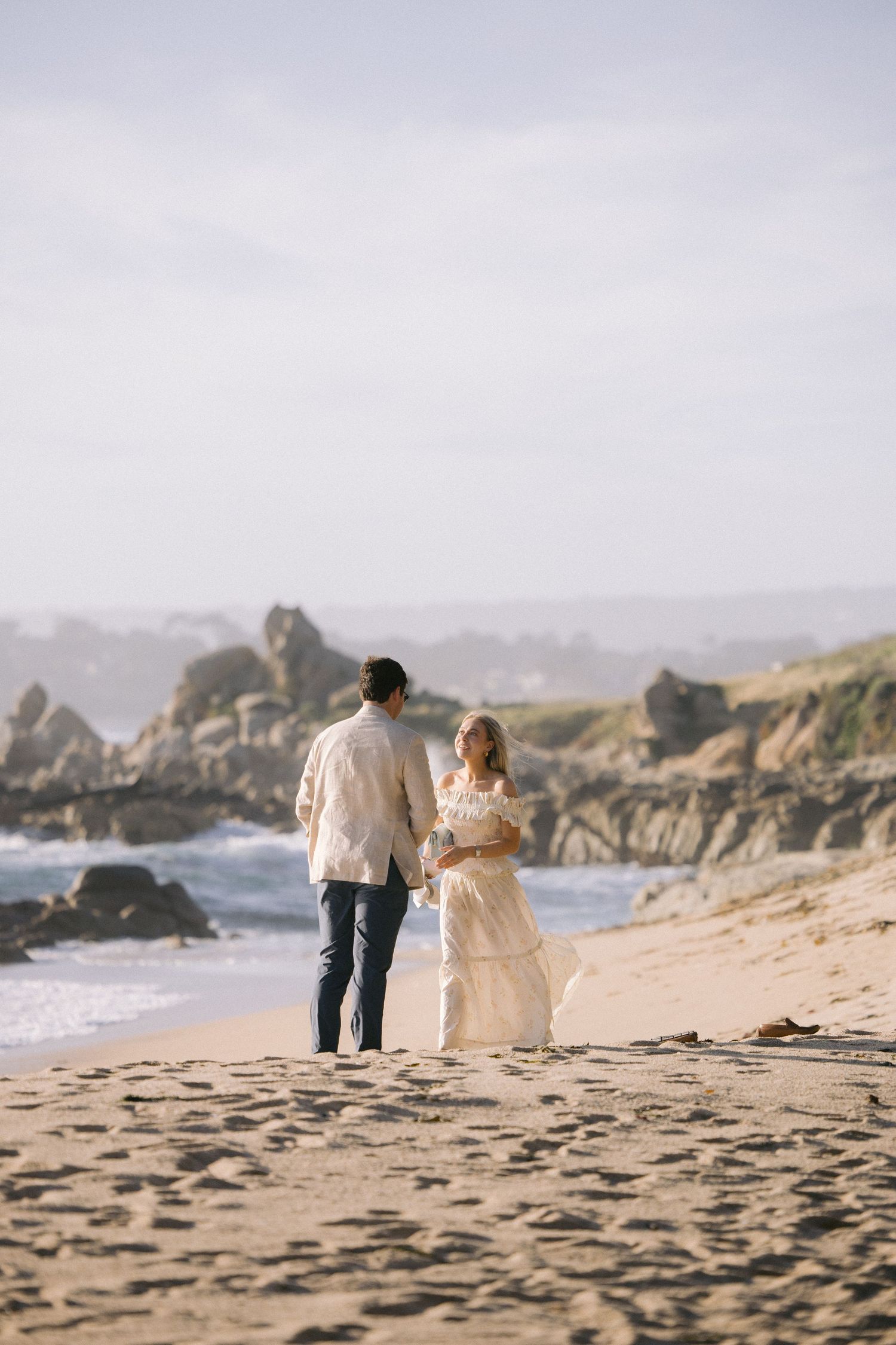 A couple stands together on a sandy beach with dramatic rocky coastline and crashing waves in the background.
