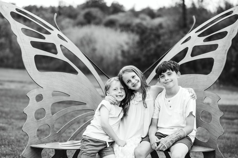 Black and white photo of family sitting on decorative butterfly bench in nature setting.