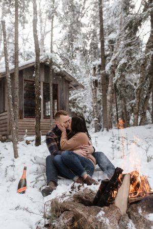 Romantic winter scene with couple cuddling by campfire outside snow-covered cabin.