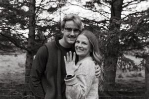 A happy couple showing off an engagement ring in a black and white forest portrait.