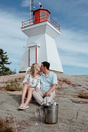 A couple shares a romantic moment near a white lighthouse with red trim on rocky terrain.
