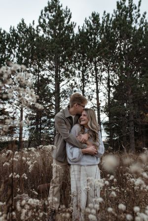 Couple embracing in a field of dried grass with evergreen trees creating a natural backdrop.
