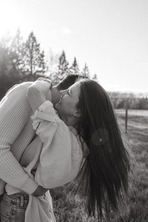 A candid black and white photo of a couple embracing in a field with sunlight and trees in the background.