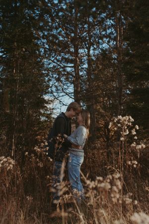 A romantic couple embraces in a moody autumn field with tall grass and evergreen trees at sunset.