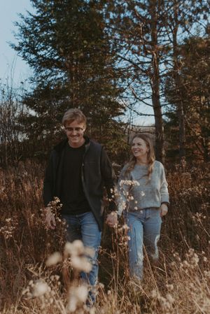 A couple strolls together through dry grass and pine trees on a fall afternoon.