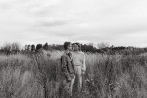 Two people standing together in a field of tall grass with pine trees in the background.