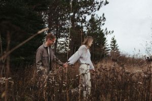 Couple walking hand in hand through tall grass with pine trees in the background.