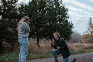 A surprise proposal moment captured outdoors on a path with evergreen trees in the background.