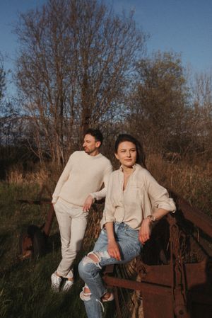 A stylish couple in casual wear leaning against rustic farm equipment in warm afternoon light.