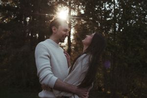 A couple shares an intimate moment outdoors with sunburst through trees in background.