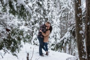 Winter engagement photo of couple embracing in snow-covered forest.