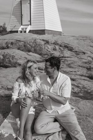 Black and white portrait of a couple sitting on rocks near a lighthouse.