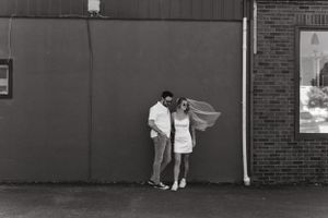 Black and white photo of couple standing against industrial wall outdoors.