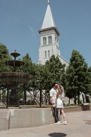 Couple poses by fountain with white church steeple in background on sunny day.