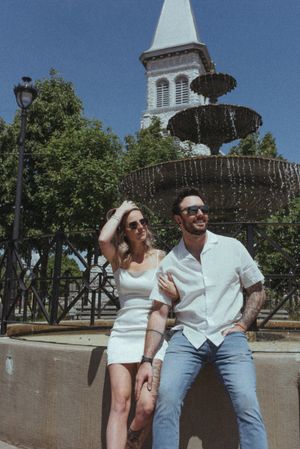 A stylish couple relaxes by a fountain with a church steeple in the background on a sunny day.