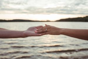 Two hands reach toward each other displaying an engagement ring against a sunset lake backdrop.