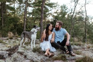 A couple relaxes on a rocky hillside with their dog against a backdrop of tall pine trees.