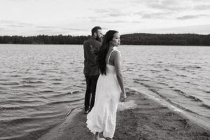 A couple stands together on the shoreline of a lake during sunset in black and white.
