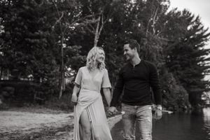 A couple strolling along a lakeside path surrounded by pine trees in black and white.