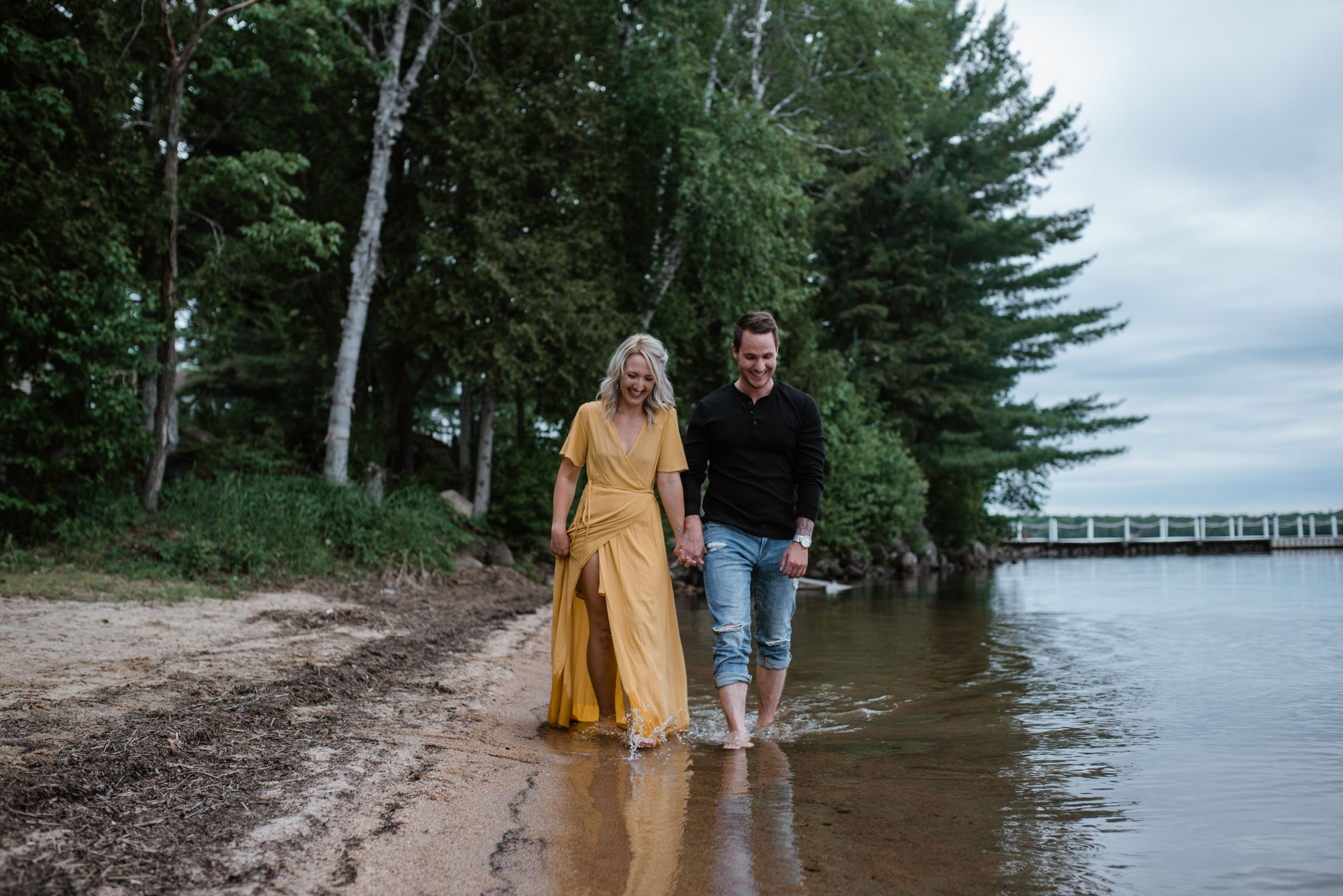 A couple walking barefoot in the shallow water along a tree-lined beach.