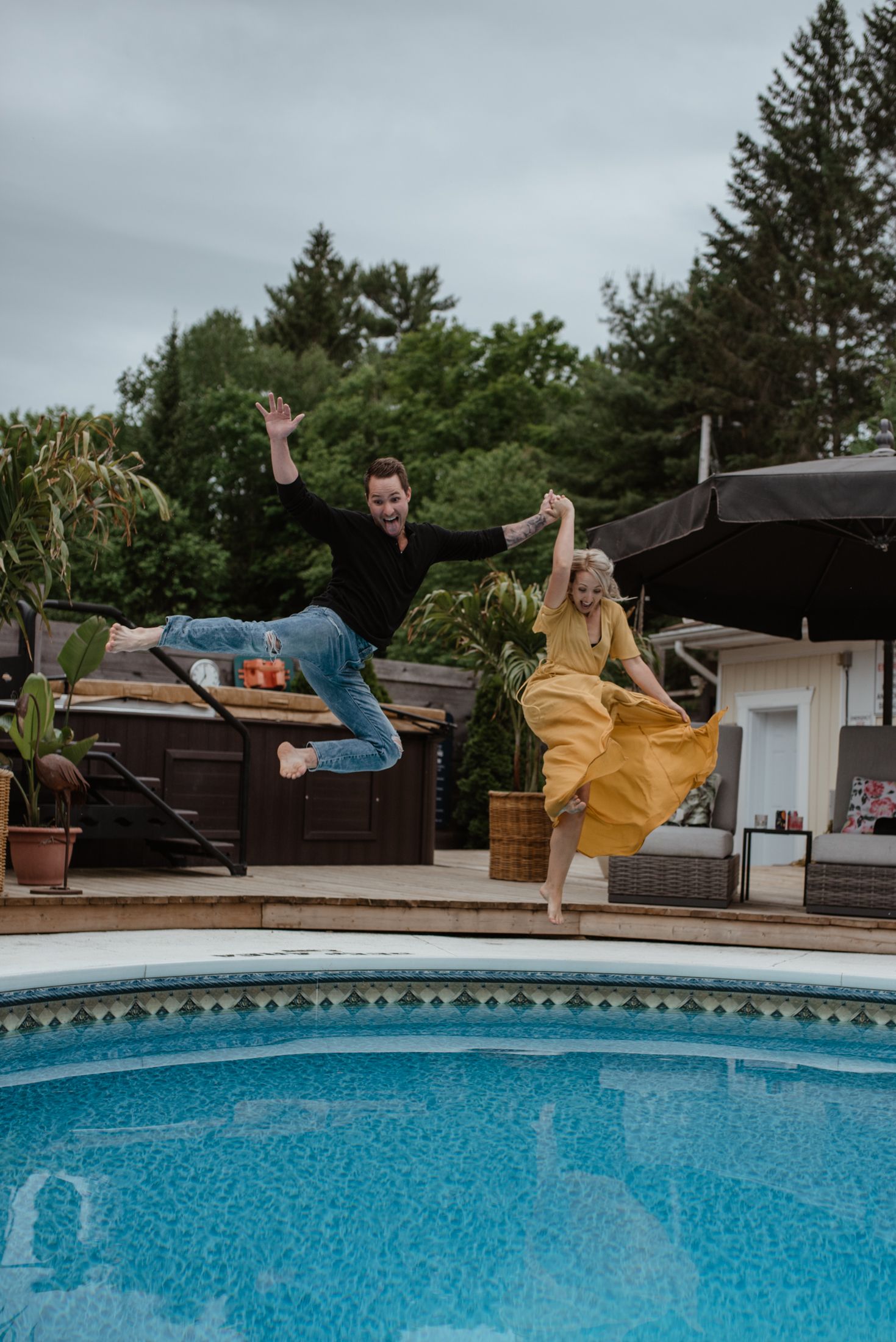 Two people joyfully jumping into a swimming pool wearing casual outfits on a cloudy day.