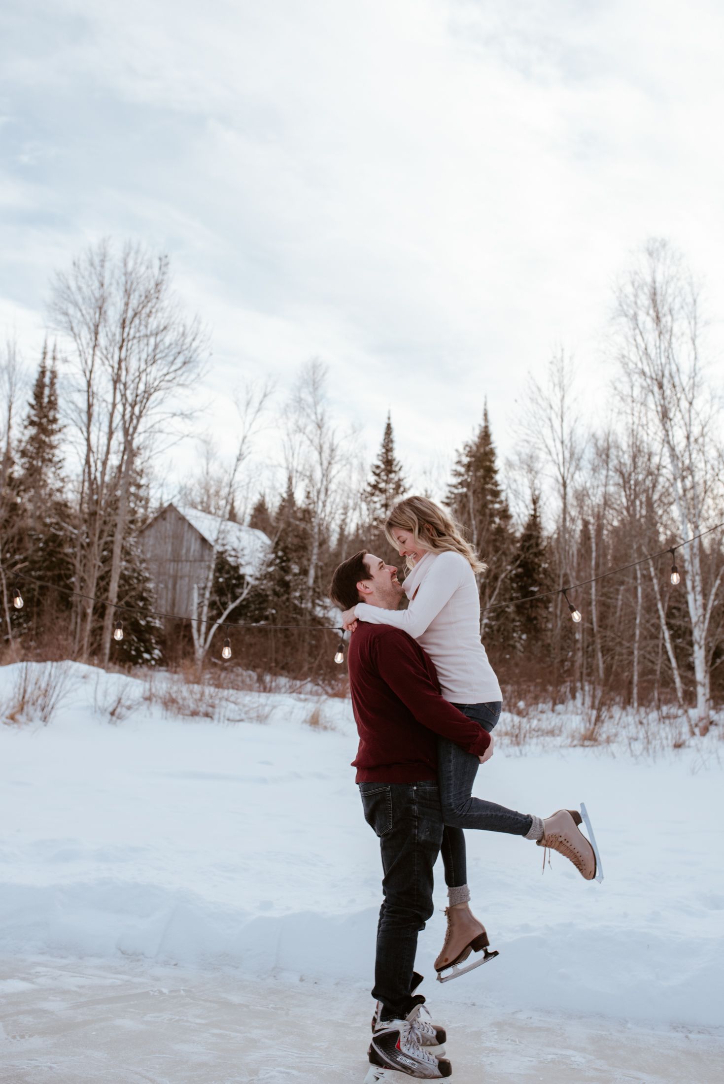 A couple embraces while ice skating in a snowy winter landscape with evergreen trees and a cabin in the background.