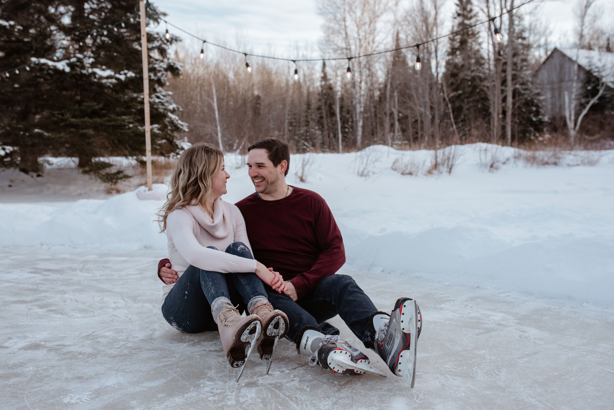 A couple sits together on ice while wearing ice skates in a snowy winter setting surrounded by trees.