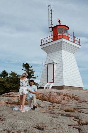 A couple poses together on rocky terrain in front of a white and red lighthouse.