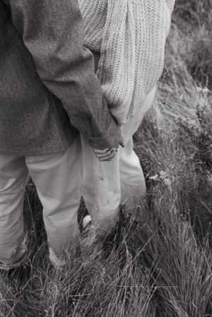 A black and white close-up of two people holding hands while standing in tall grass.