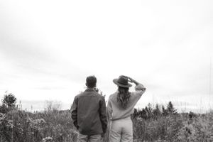 A couple stands together in a field of tall grass against an overcast sky.
