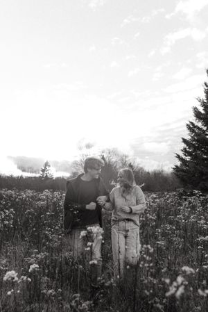 Black and white photo of a couple standing among wildflowers in a meadow with evergreens.