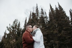 A couple embracing in front of snow-dusted evergreen trees on a winter day.