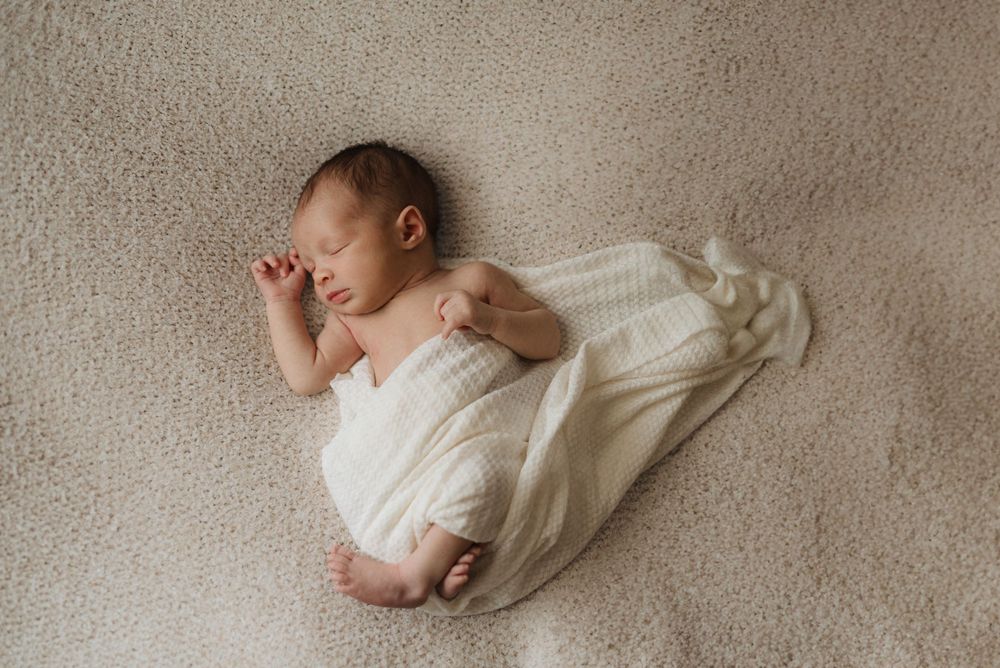 Newborn baby peacefully sleeping on a soft beige blanket, wrapped in a white swaddle.