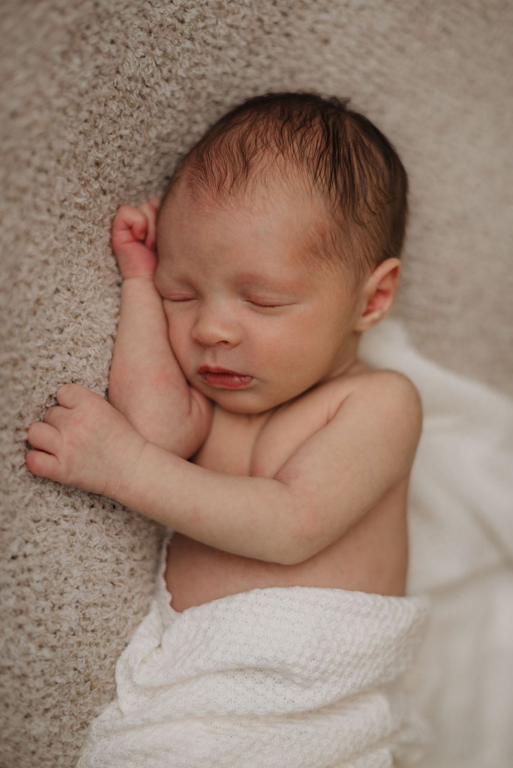Newborn baby peacefully sleeping on a soft beige blanket, wrapped in a white swaddle.