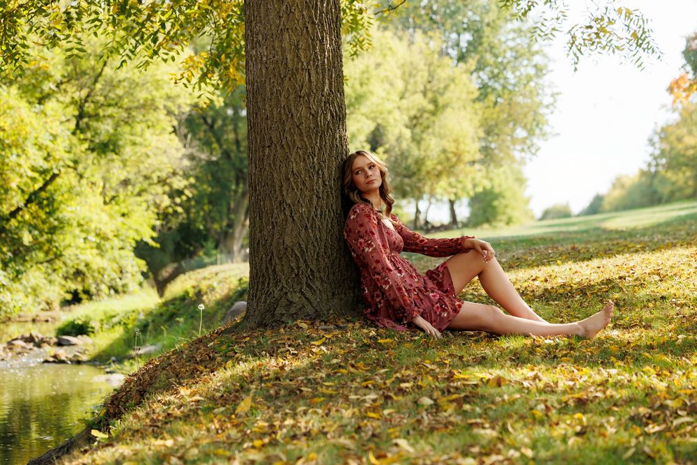 A person in a burgundy dress sits beneath a large tree on an autumn day as golden sunlight filters through leaves.