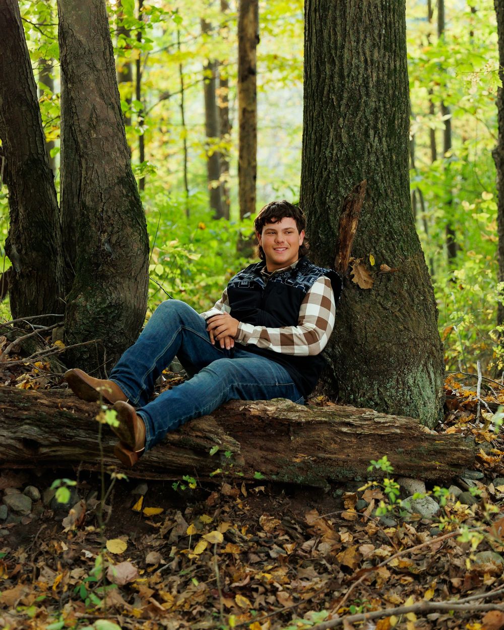 A person in jeans and plaid shirt reclines against a fallen log in an autumn forest with golden leaves on the ground.