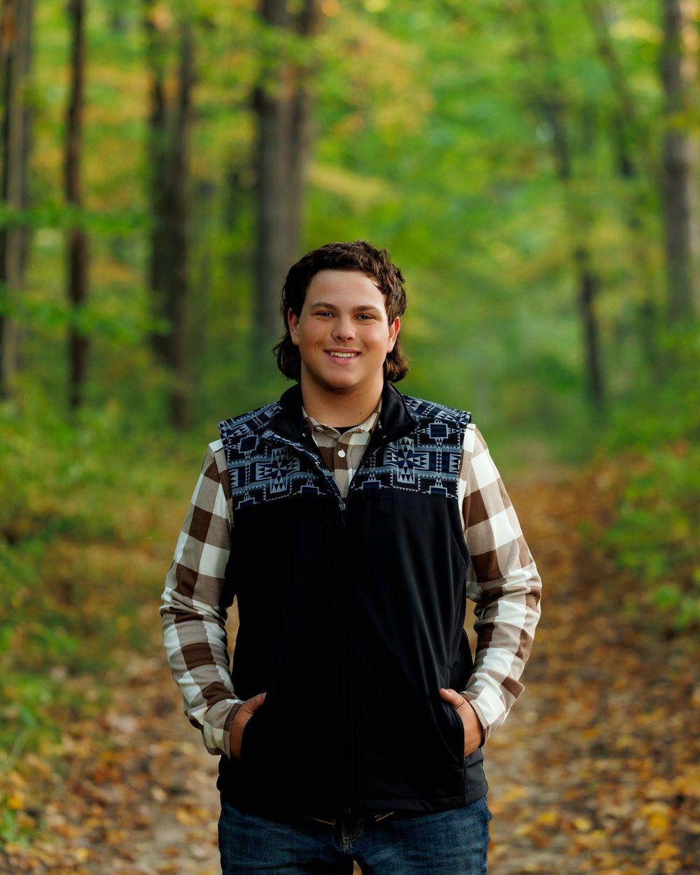 Person wearing a black vest and plaid shirt poses outdoors among autumn trees with fallen leaves on the ground.