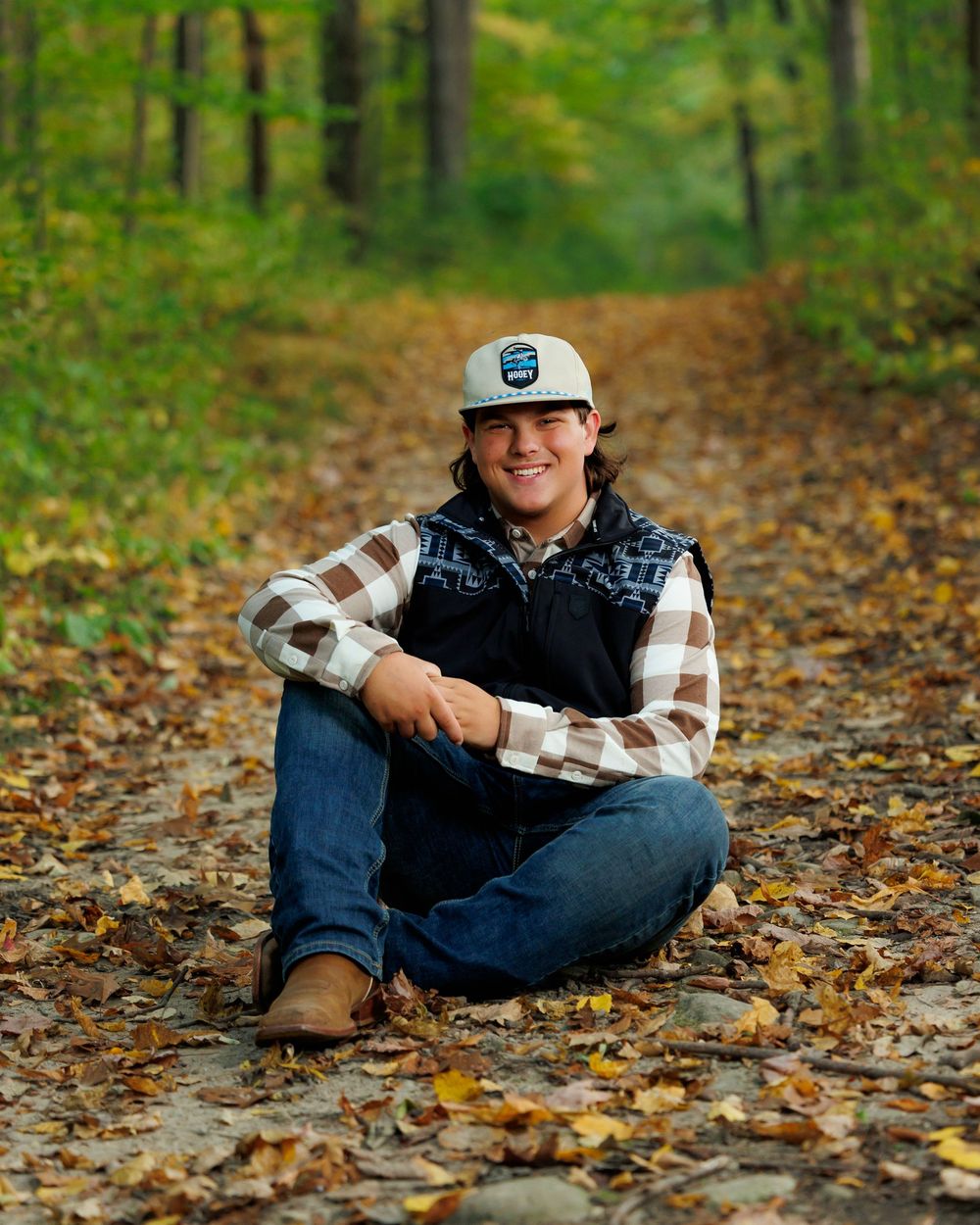 A figure sits on a leaf-covered forest trail wearing a plaid shirt and baseball cap on an autumn day.