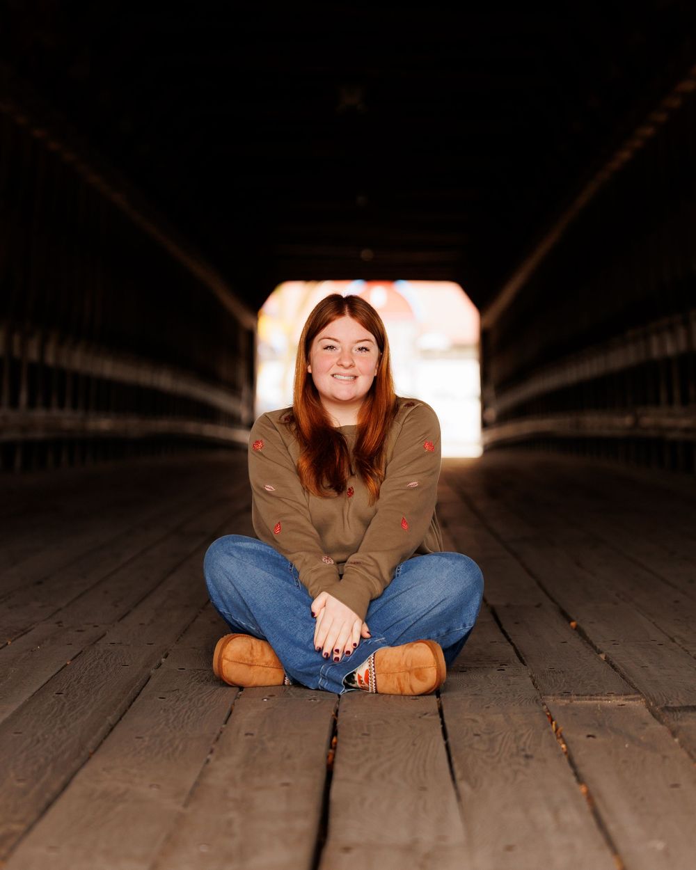 Person sits cross-legged on wooden bridge floor with light visible at tunnel's end, wearing brown sweater and jeans.